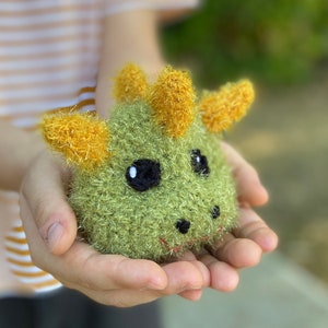 May include: A handmade, green and yellow crocheted creature with black eyes and a red mouth. The plush toy has yellow horns and is held in open hands. The toy is a whimsical, soft sculpture.