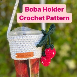 May include: White crocheted boba holder with a brown leather tag that says "Hand Made". The holder has a green crocheted strawberry charm attached to it.