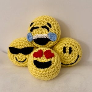 May include: Four yellow crocheted emoji faces. One has sunglasses, another has red heart eyes, a third has a smiling face, and the last one is laughing with blue tears. The emojis are handmade and have black and white details.