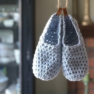 May include: A pair of light blue crocheted slippers hanging from a natural twine. The slippers have a textured stitch pattern and are suspended by small brown tags. The background is blurred, suggesting an indoor setting.