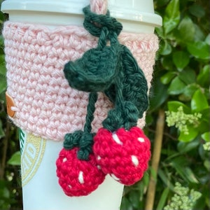 May include: A pink crocheted coffee cup cozy with green leaves and red strawberries attached.
