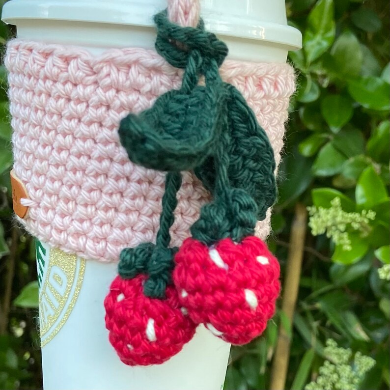 May include: A pink crocheted coffee cup cozy with green leaves and red strawberries attached.