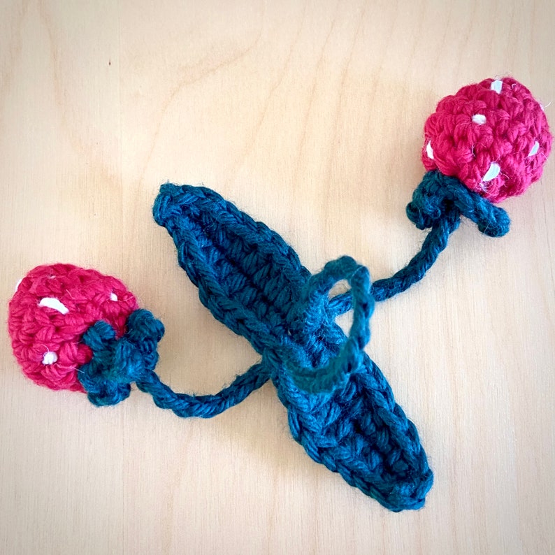 May include: A crocheted strawberry garland with two red strawberries and a green leafy stem. The garland is made of dark blue yarn.