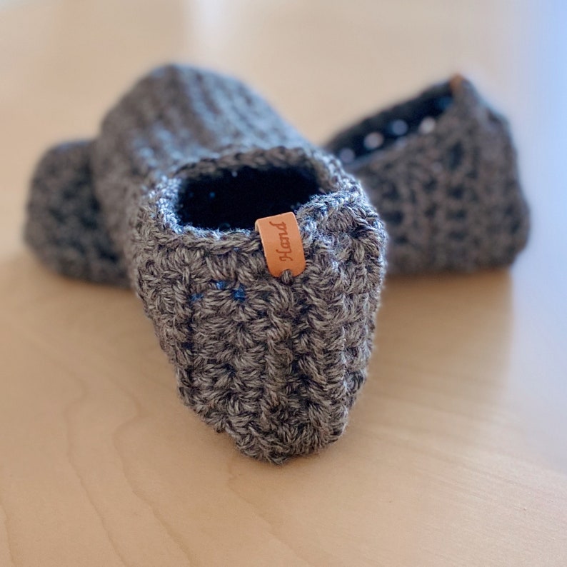 May include: A pair of gray crocheted slippers with a brown leather tag that says "handmade".