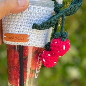 May include: White crocheted cup cozy with a brown "Hand Made" label and a green crocheted strawberry charm with white dots.