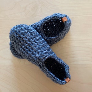 May include: A pair of gray crocheted slippers with brown leather tags. The slippers are made with a textured stitch and have a simple, comfortable design.