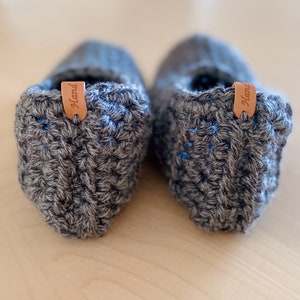 May include: A pair of gray crocheted slippers with blue accents and a brown leather tag that says "Hand" on each slipper.