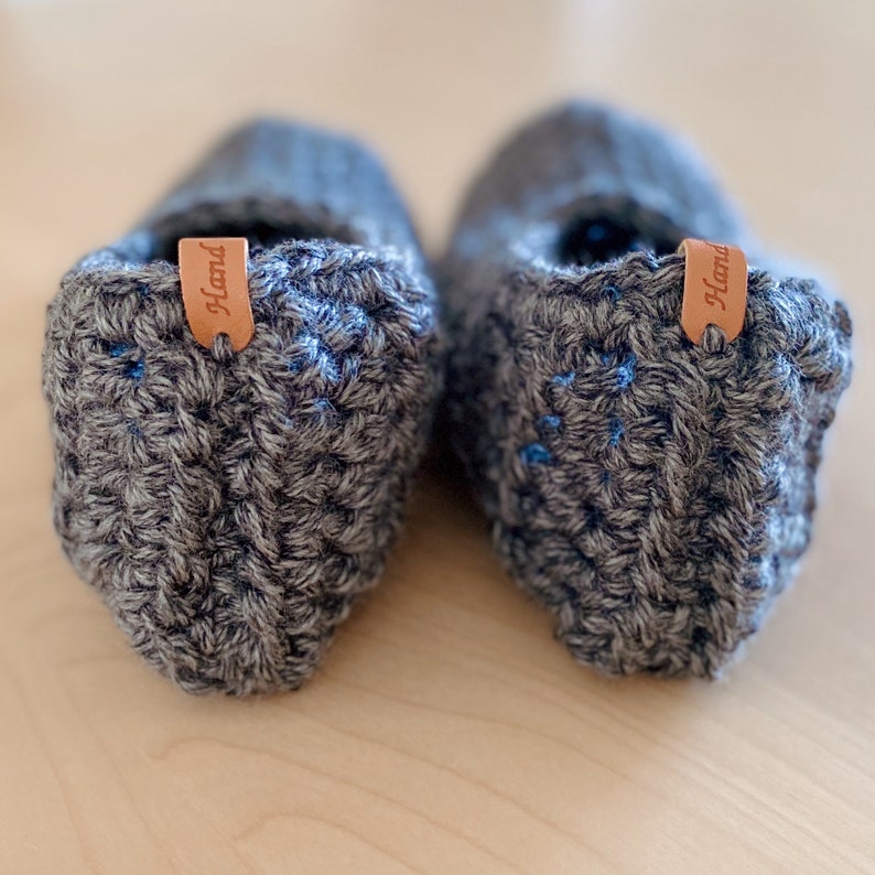 May include: A pair of gray crocheted slippers with blue accents and a brown leather tag that says "Hand" on each slipper.
