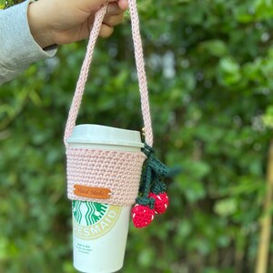 May include: Pink crocheted coffee cup holder with a strap, featuring a small crocheted strawberry design. The holder has a brown tag that says "Hand Made".
