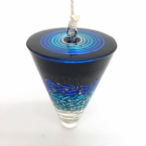 May include: A pendulum with a black, cone-shaped body. The top features concentric blue rings, while the lower section is filled with blue and teal beads. A silver loop and white cord are attached for hanging.