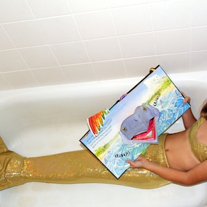 May include: A young person in a bathtub wearing a gold mermaid tail and bikini top, holding a colorful illustrated book. The book features a cartoon hippo with the word "CRUNCH!" on the page. Blue swim goggles are on their head.