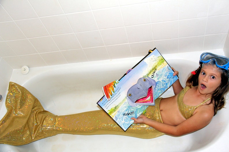 May include: A young person in a bathtub wearing a gold mermaid tail and bikini top, holding a colorful illustrated book. The book features a cartoon hippo with the word "CRUNCH!" on the page. Blue swim goggles are on their head.