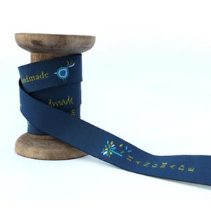 Puede incluir: Un carrete de cinta azul oscuro con el texto "Handmade" en letras amarillas y un pequeño diseño de pájaro azul. La cinta está en un carrete de madera.