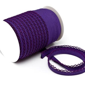 Könnte beinhalten: Eine Rolle lila Band mit dekorativem Rand. Das Band hat eine tiefe violette Farbe und eine strukturierte, gewebte Optik. Ein Stück des Bandes ist abgerollt und zeigt das komplizierte Design.