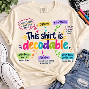 May include: A cream-coloured short-sleeve t-shirt featuring the text This shirt is decodable in large, colourful letters. Surrounding the text are various educational annotations with arrows pointing to specific linguistic concepts like Closed Syllable, Digraphs, r-controlled, Latin-based Prefix, Silent-e, and Latin-based Suffix -able.