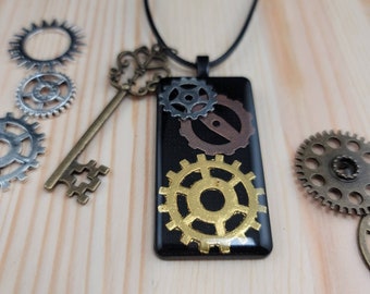 Op de afbeelding: Een zwarte rechthoekige hangerketting met gouden, zilveren en koperen tandwielen op een zwarte achtergrond. De ketting is aan een zwart koord.