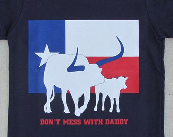 May include: A black t-shirt with a white silhouette of a Texas longhorn and calf standing in front of a Texas flag. The text "Don't mess with Daddy" is printed below the image.