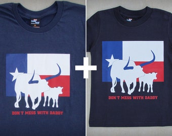 May include: Two navy blue t-shirts with a Texas flag design. The larger shirt features a white silhouette of a longhorn steer and calf with the text "Don't Mess With Daddy". The smaller shirt features the same design but with a smaller silhouette of the steer and calf.