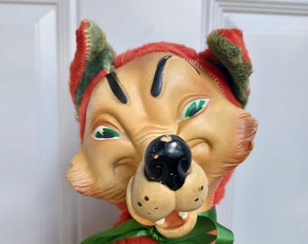 Puede incluir: Un juguete de peluche vintage en forma de zorro con un cuerpo estampado en rojo y verde, ojos verdes y nariz negra. El zorro lleva una cinta verde alrededor del cuello.