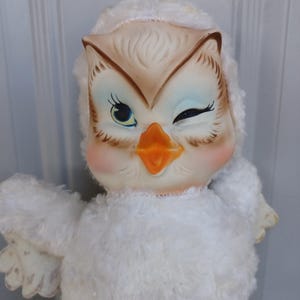Puede incluir: Una mu&ntilde;eca de b&uacute;ho vintage con una expresi&oacute;n gui&ntilde;ando un ojo. El b&uacute;ho tiene una cara color crema con detalles en marr&oacute;n y azul, un pico naranja y mejillas rosadas. Su cuerpo est&aacute; cubierto de material blanco y esponjoso, y tiene peque&ntilde;as alas extendidas.