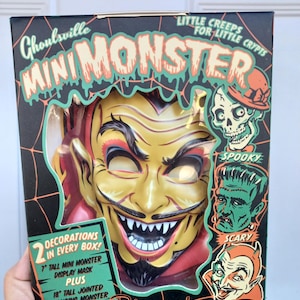May include: A Ghoulsville Mini Monster display mask in a black box with green text. The box features a yellow devil mask, illustrations of a skull, Frankenstein, and a devil, with text stating 2 decorations in every box, 7 inch mask, and 18 inch monster.