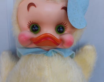 May include: A vintage plush duck doll with a plastic face. The doll has a yellow body, a light blue bonnet, and a light blue neck ribbon. The face is peach-colored with green eyes, rosy cheeks, and an orange beak. The doll has a fluffy, soft texture.