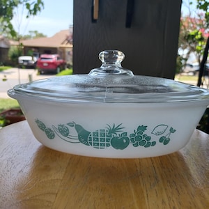 May include: An oval white glass casserole dish with a clear glass lid and a green fruit pattern featuring a pineapple, grapes, and pears printed along the side. The dish has integrated handles on both ends and a round knob on the lid.