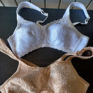 May include: Two bras, one white and one beige, are displayed. Both bras feature a delicate lace pattern across the cups and adjustable straps. The white bra has a matching lace trim, while the beige bra has a smooth, solid trim.