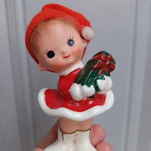 Puede incluir: Una figura navideña vintage de una niña con un vestido y sombrero rojos, sosteniendo un regalo verde con un lazo rojo. La figura tiene una falda y botas blancas. La niña tiene un ojo azul y un ojo negro.