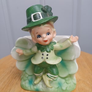 May include: A ceramic figurine of a leprechaun wearing a green hat and suit, standing in front of a white and green decorative base. The figurine has a smiling face with blue eyes and red lips. The base has a leaf-like design.