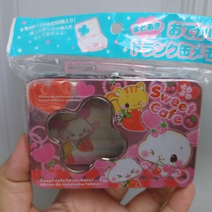 May include: A rectangular metal tin with a clover-shaped cutout window, featuring pink and red colors, cartoon animal illustrations, and the text "Sweet Cafe". The tin is packaged in a clear plastic bag with a blue header card containing Japanese text.