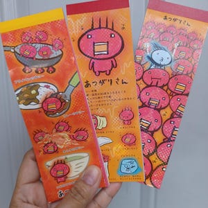 May include: Three rectangular paper memo pads featuring the character Atsugari-san. The designs display the pink, spiky-haired character in various scenarios, including cooking in a pan, eating curry, and sweating in a crowd, with Japanese text.