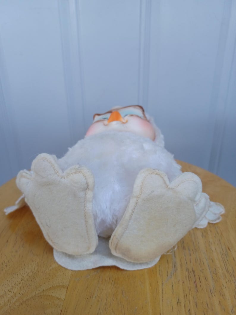 Puede incluir: Un juguete de peluche de b&uacute;ho blanco vintage con grandes pies de fieltro. El b&uacute;ho tiene un pico de color naranja claro, ojos azules y un cuerpo suave y esponjoso. El b&uacute;ho est&aacute; acostado sobre su espalda sobre una superficie de madera.