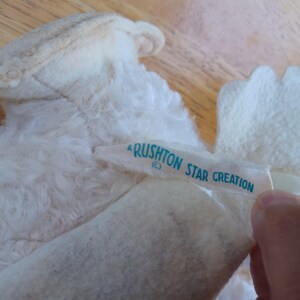 Puede incluir: Primer plano de un juguete de peluche blanco vintage con una etiqueta que dice "Rushton Star Creation" en texto azul claro. El juguete tiene una textura suave y esponjosa y es parcialmente visible.