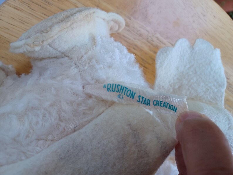 Puede incluir: Primer plano de un juguete de peluche blanco vintage con una etiqueta que dice "Rushton Star Creation" en texto azul claro. El juguete tiene una textura suave y esponjosa y es parcialmente visible.