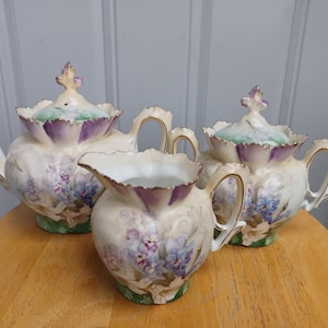 May include: A vintage porcelain tea set with a teapot, sugar bowl, and creamer. The set features a floral design in shades of purple and blue on a cream background. The teapot and sugar bowl have lids with decorative finials.
