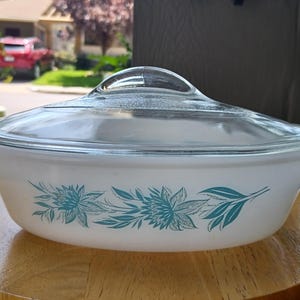 May include: An oval white glass casserole dish with a clear glass lid. The exterior of the dish features a repeating teal floral pattern consisting of stylised flowers and leaves arranged in a horizontal band around the centre of the vessel.