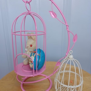 Può includere: Un espositore decorativo in metallo rosa a forma di gabbia per uccelli, con un piccolo coniglietto di peluche bianco e un uovo di Pasqua blu all'interno. È presente anche una gabbia bianca più piccola. Ideale per le decorazioni pasquali.