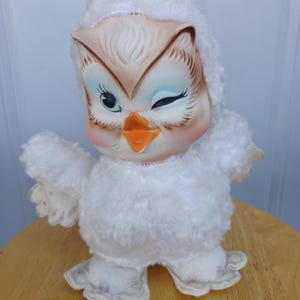 Puede incluir: Un juguete de peluche vintage con forma de b&uacute;ho con un gui&ntilde;o. El b&uacute;ho tiene una cabeza y un cuerpo de color crema, con una capucha blanca y esponjosa. Su cara presenta ojos azules, mejillas rosadas y un pico naranja. El b&uacute;ho est&aacute; sobre una superficie de madera.