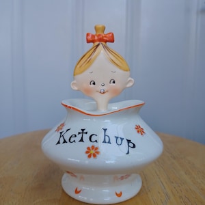May include: A white ceramic ketchup server featuring a whimsical character topper with blonde hair and an orange bow. The base is decorated with small orange flower motifs and the word Ketchup written in black script across the front surface.