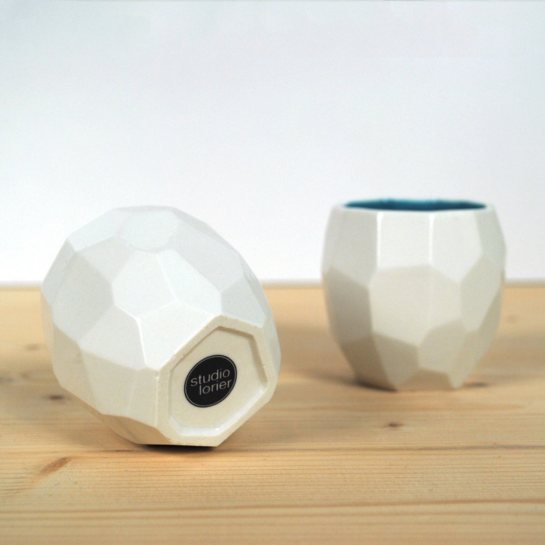May include: Two white ceramic cups with a geometric design. The cup on the left has a black sticker with the text "studio lorier".