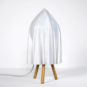 Puede incluir: Una lámpara blanca con una pantalla de tela y una base de trípode de madera.