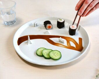 May include: A white plate with a brown sauce design resembling a river. Three pieces of sushi are arranged on the plate, along with cucumber slices. A pair of chopsticks is holding a piece of sushi.