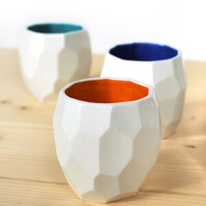 May include: Three geometric ceramic cups with white exteriors and different colored interiors. The cups are teal, blue, and orange.