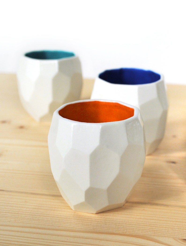 May include: Three geometric ceramic cups with white exteriors and different colored interiors. The cups are teal, blue, and orange.