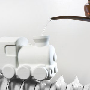 Puede incluir: Una maceta blanca con forma de tren está sobre un radiador. El agua se vierte de una regadera de cobre a la maceta. El tren tiene un cuerpo rectangular con ruedas circulares y una chimenea.