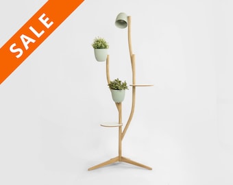 May include: A wooden plant stand with a light fixture and three shelves. The stand is shaped like a tree with branches and a base. The shelves are white and the plant pots are light green.