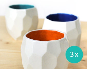May include: Three white ceramic cups with geometric designs. The cups have different colored interiors: teal, blue, and orange.