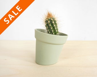 May include: A small green ceramic pot with a cactus plant inside. The pot is tilted to the side and the cactus is leaning out of the pot.