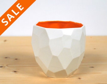 May include: White ceramic planter with a geometric design and an orange interior. The planter is sitting on a light wood surface.